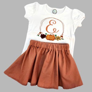 Könnte beinhalten: Ein weißes Kurzarmshirt mit Puffärmeln und Rundhalsausschnitt, verziert mit einem orangefarbenen Buchstaben E, einem Kürbis und Herbstblättern. Dazu ein einfarbiger, rostfarbener Rock mit elastischem Bund und gerafftem Stoff.