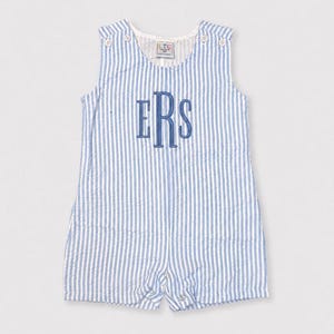 Peut inclure: Une barboteuse sans manches en seersucker avec un motif à rayures verticales bleu clair et blanc. Le vêtement présente une encolure ronde, quatre boutons blancs aux épaules et un monogramme bleu brodé avec les lettres ERS sur la poitrine.