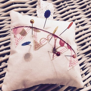 May include: A white pincushion with a pink and brown fabric design. The pincushion is filled with pins with colourful heads.