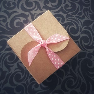 Puede incluir: Una pequeña caja de regalo cuadrada de papel kraft con una cinta rosa de lunares atada con un lazo. La caja tiene recortes redondeados en los lados. El fondo es una superficie oscura y azul con dibujos.