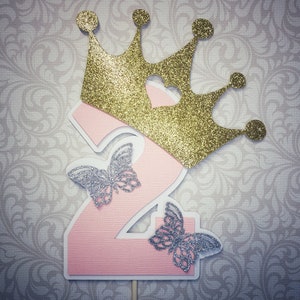 May include: Pink number two cake topper with a gold glitter crown and two silver glitter butterflies.
