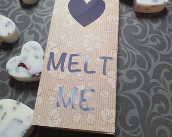May include: A box with a heart-shaped cutout and the words "MELT ME" printed on it. The box is surrounded by small white heart-shaped wax melts with dried flowers and gold flakes.