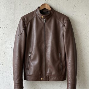 Puede incluir: Chaqueta de cuero marrón con cuello alzado y cierre de cremallera. La chaqueta tiene dos bolsillos con cremallera en el pecho y dos en los laterales. Cuelga de una percha de madera sobre un fondo neutro.