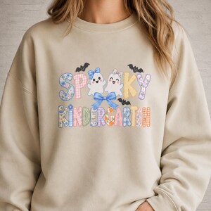 May include: A beige crewneck sweatshirt featuring a graphic with the text "SPOOKY KINDERGARTEN" in colourful, patterned block letters. The design includes two white ghosts with bows, a large blue bow, and small black bats scattered around.