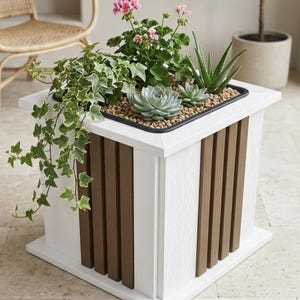 May include: A square white and brown planter box with a variety of plants, including pink flowers, succulents, and ivy. The planter has vertical brown slats and is placed on a tiled floor. The plants are in a black tray.