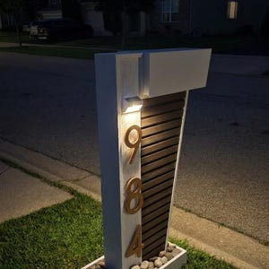 May include: A vertical white wooden post with a dark brown slatted panel and the gold house numbers 9, 8, and 4 mounted on the front. A small light fixture is attached above the numbers, and the base is filled with smooth gray river stones.