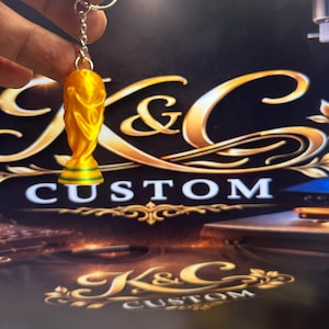 May include: A bright yellow, three-dimensional trophy-shaped keychain featuring a smooth, curved silhouette. The base of the trophy is accented with a thin horizontal green stripe. The keychain is attached to a standard silver metal split ring.