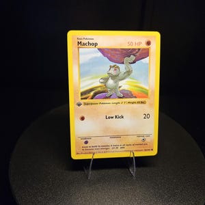 May include: A Machop trading card with a yellow border, featuring a gray creature lifting a large purple rock. The card displays 50 HP, the move Low Kick with 20 damage, and text including Superpower Pokémon, Length 2' 3", and Weight 43 lbs.