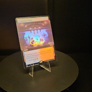 Peut inclure: Une carte à collectionner Magic: The Gathering rectangulaire intitulée Training Center, présentant une carte de terrain avec une finition holographique. L'illustration montre une structure lumineuse bleue et orange avec du texte de jeu.