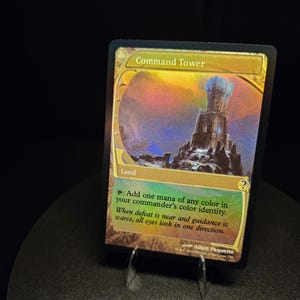 Può includere: Una carta collezionabile olografica di Magic: The Gathering intitolata Command Tower. La carta presenta un'illustrazione di una torre in pietra, il testo Land e l'abilità di aggiungere un mana di qualsiasi colore. Include testo narrativo e nome dell'artista.