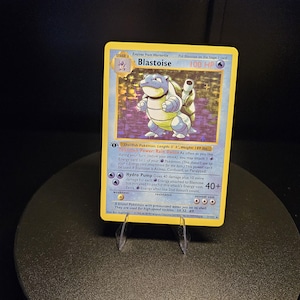 May include: A Blastoise trading card featuring a blue turtle-like creature with cannons on its shell. The card displays 100 HP, the Rain Dance Pokémon Power, and the Hydro Pump attack. It includes text detailing the Pokémon's length and weight.
