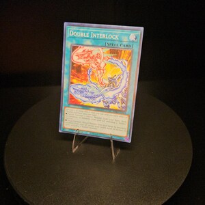 May include: A rectangular trading card titled Double Interlock, identified as a Spell Card. The card features a vibrant illustration with swirling blue and orange energy patterns, encased in a protective plastic sleeve and displayed on a clear stand.