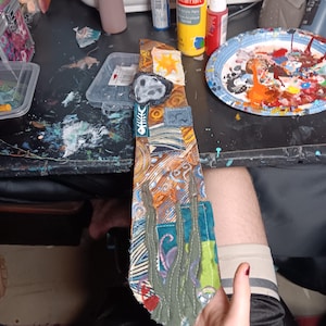 May include: A necktie featuring a patchwork design with various fabric patterns in shades of orange, brown, and blue. The tie includes appliqued elements such as a white fish skeleton, a grey moon shape, and green seaweed-like fabric strips.
