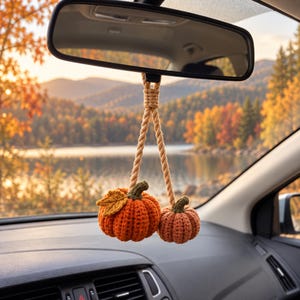 May include: A hanging car accessory featuring two crocheted pumpkins in shades of orange, suspended from a twisted beige rope. The larger pumpkin includes a single crocheted leaf, and both pumpkins have small, dark green crocheted stems.