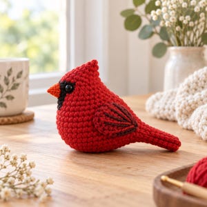 May include: A small, handcrafted crochet cardinal bird figurine. The bird is primarily bright red with a black patch around its dark, round eye and a small orange beak. It features a pointed crest on its head and dark brown stitched wing details.
