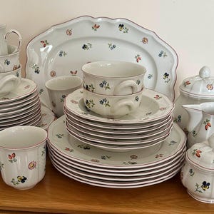 Op de afbeelding: Een wit keramieken servies met een delicaat bloemmotief van kleine, kleurrijke bloemen. De set bestaat uit gestapelde borden, kommen met oren, mokken, een theepot met deksel, een suikerpot met deksel, een melkkan en een grote ovale serveerschaal.