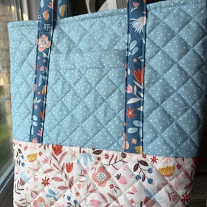 Peut inclure: Un sac fourre-tout matelassé composé d'un tissu bleu clair à pois blancs sur la partie supérieure et d'un tissu crème à motifs floraux multicolores sur la partie inférieure. Le sac possède deux anses assorties en tissu floral bleu.