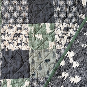 May include: A quilted patchwork blanket featuring a variety of fabric squares with forest-themed prints. The patterns include silhouettes of deer, bears, and pine trees in shades of forest green, charcoal grey, black, and cream with diamond stitching.