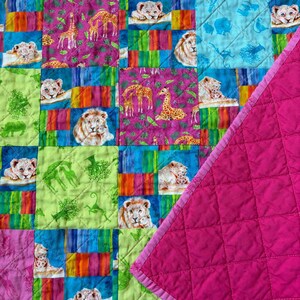 May include: A quilted blanket featuring a patchwork of squares with vibrant prints of lions, giraffes, and rhinoceroses in shades of pink, lime green, and blue, with a solid hot pink quilted fabric folded over the corner showing diamond stitching.