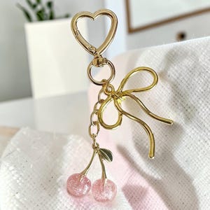 May include: A gold-toned metal keychain featuring a heart-shaped clasp, a decorative bow, and two dangling pink translucent cherry charms with glitter accents. The cherries are attached to the main ring by a short chain and a small green leaf.