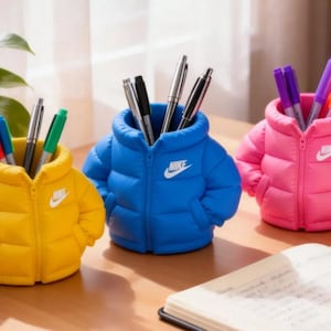 Puede incluir: Tres portalápices con forma de chaqueta acolchada en amarillo, azul y rosa, cada uno con un logotipo de Nike blanco en el pecho. Estos organizadores de escritorio están diseñados para guardar bolígrafos y rotuladores, imitando el acolchado.