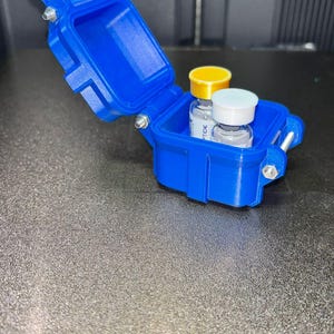 Puede incluir: Un pequeño estuche de almacenamiento rectangular de color azul brillante con un mecanismo de bisagra y cierre. La tapa está abierta, revelando dos viales de vidrio en su interior. Un vial tiene una tapa amarilla y el otro una blanca. Bisagras metálicas.