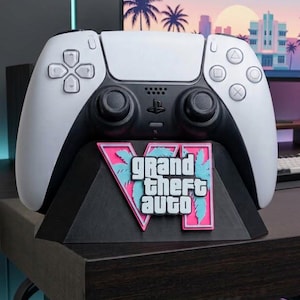 Op de afbeelding: Een witte en zwarte draadloze gamecontroller rustend op een zwarte driehoekige standaard. De standaard is voorzien van een roze en lichtblauw logo met de tekst "GRAND THEFT AUTO VI" in witte letters, met palmbomsilhouetten achter het Romeinse cijfer.