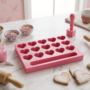 Puede incluir: Un molde rectangular de madera rosa con quince cavidades en forma de corazón y una ranura rectangular larga. El conjunto incluye un sello rosa a juego en forma de corazón con mango, cuencos con virutas de colores, un tarro de glaseado y rodillos.