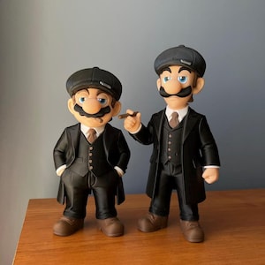 Puede incluir: Dos figuras de personajes de Mario vestidos con trajes negros, sombreros y zapatos marrones. Una figura sostiene un cigarro. Las figuras tienen bigotes y están de pie sobre una superficie de madera. Una versión única de los personajes clásicos de videojuegos.
