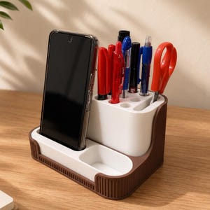 Pode incluir: Um organizador de secretária bicolor com interior branco e exterior castanho, apresentando uma ranhura vertical para um smartphone, um tabuleiro retangular e vários compartimentos cilíndricos com várias canetas, marcadores e uma tesoura.
