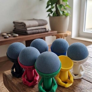 May include: A collection of seven small, stylised figures in black, red, green, yellow, and white, each featuring a seated posture with legs crossed. Each figure supports a round, blue, fabric-covered smart speaker device on its head.