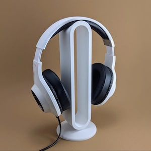 May include: A pair of white over-ear headphones with black ear cushions resting on a white vertical oval-shaped headphone stand. The headphones feature a white headband and a black audio cable extending from the bottom of the left ear cup.
