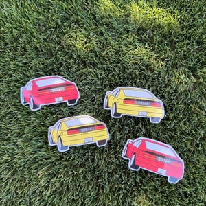 May include: Four die-cut stickers depicting the rear view of a sports car. Two stickers are red and two are yellow. Each sticker features a black rear light bar with the text 300zx in white lettering, along with a small white license plate area.