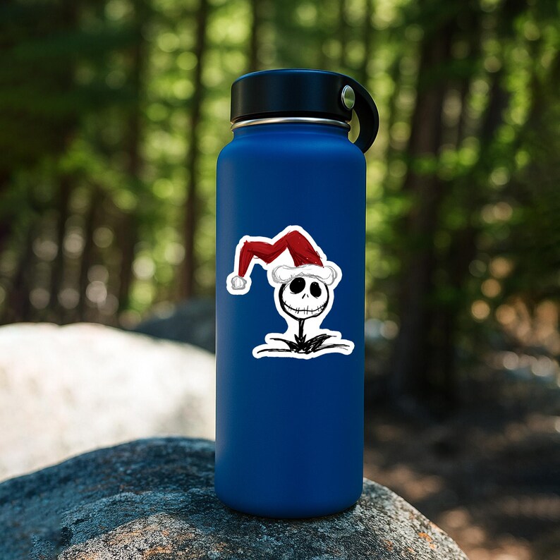 May include: A matte dark blue water bottle featuring a sticker of a white skeletal character head wearing a red and white Santa hat. The character has large hollow eyes and a stitched mouth, rendered in a sketch-like black ink style.