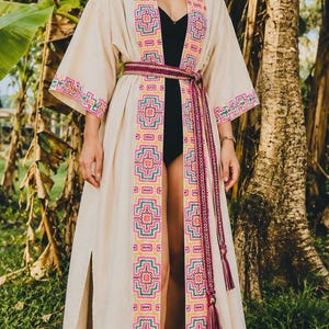 May include: A long, cream-colored kimono-style robe with vibrant embroidered details in pink, green, and yellow. The robe features a matching belt and tassel, and is worn over a black garment. The robe is open in the front, revealing the legs.