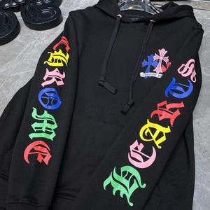 May include: Black hoodie with colorful "Chrome Hearts" lettering on the sleeves. The hoodie features a drawstring hood and a small emblem with a cross design on the chest. The lettering and emblem are in various colors.