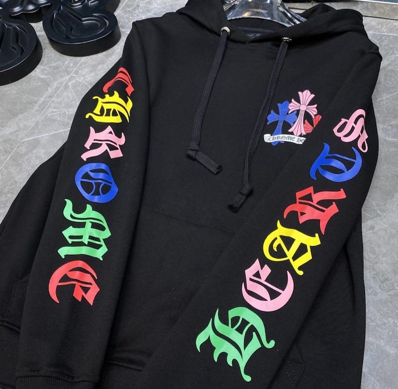 May include: Black hoodie with colorful "Chrome Hearts" lettering on the sleeves. The hoodie features a drawstring hood and a small emblem with a cross design on the chest. The lettering and emblem are in various colors.