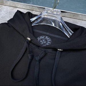 May include: Black hooded sweatshirt with drawstrings and silver-toned metal accents. The sweatshirt features a circular emblem with a floral design and text. The garment is hanging on a clear plastic hanger.