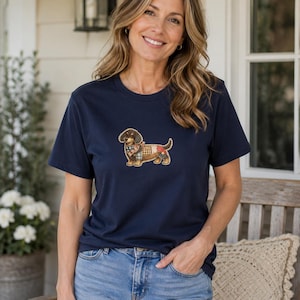 Può includere: Una t-shirt a maniche corte blu navy con una grafica centrale raffigurante un cane bassotto. Il cane è rappresentato con un motivo a patchwork in vari colori, tra cui marrone, rosso, bianco e blu, e indossa un piccolo foulard a fantasia.