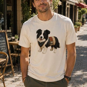 Può includere: Una t-shirt a maniche corte color crema con un'illustrazione grafica di un cane border collie bianco e nero. Il cane indossa una bandana blu scuro a motivi intorno al collo e un maglione patchwork multicolore sulla schiena.
