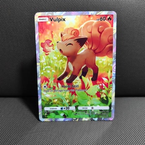 Peut inclure: Une carte à collectionner Vulpix représentant une créature brune ressemblant à un renard avec une grande queue enroulée dans un champ vert vif. La carte affiche le texte Vulpix, 60 PV et l'attaque Hold Still, qui soigne 20 dégâts de ce Pokémon.