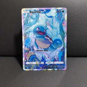 May include: A Squirtle trading card featuring a blue turtle character in an underwater scene with coral and aquatic plants. The card displays the name Squirtle, 70 HP, a water energy symbol, the Water Gun attack, and a weakness of +20.