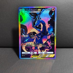May include: A holographic Charizard ex trading card featuring a dark blue dragon illustration with vibrant rainbow-colored accents. The card displays 180 HP, the attacks Slash and Crimson Storm, and a rainbow-colored border on a black background.