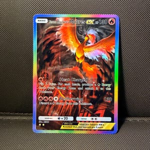 Puede incluir: Una carta coleccionable Team Rocket's Moltres ex con un borde holográfico arcoíris. La carta muestra una ilustración de un ave de fuego, 130 HP y los ataques Heat Charged y Netherwing. El texto incluye reglas del juego, debilidad y coste.
