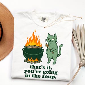 May include: A white t-shirt featuring a graphic of a green cat holding a wooden spoon next to a dark green cauldron over a fire. The text below reads, "that's it. you're going in the soup." The shirt has a Comfort Colors brand label at the neck.