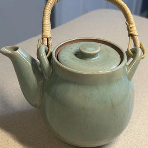 May include: A light green ceramic teapot with a glossy, speckled glaze and a rounded body. It features a matching lid with a small knob and a curved spout. A flexible, light-colored woven rattan handle is attached to two upright ceramic loops.