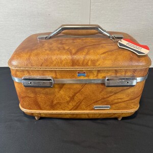 May include: A rectangular, amber-colored hardshell train case with a textured finish. It features a silver-tone metal handle, two silver-tone latches, and a small American Tourister brand tag attached to the handle. The case has four small feet.