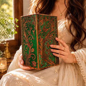 May include: A rectangular, green box with intricate, carved floral and vine designs in a contrasting brown color. The box is held by a person wearing a cream-colored dress. The box has a vintage, antique aesthetic.