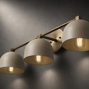 May include: A three-light wall fitting featuring a horizontal brass bar and a circular wall mount. Three dome-shaped, matte beige shades are attached to the bar, each housing a single glowing filament light bulb that casts a warm light downwards.