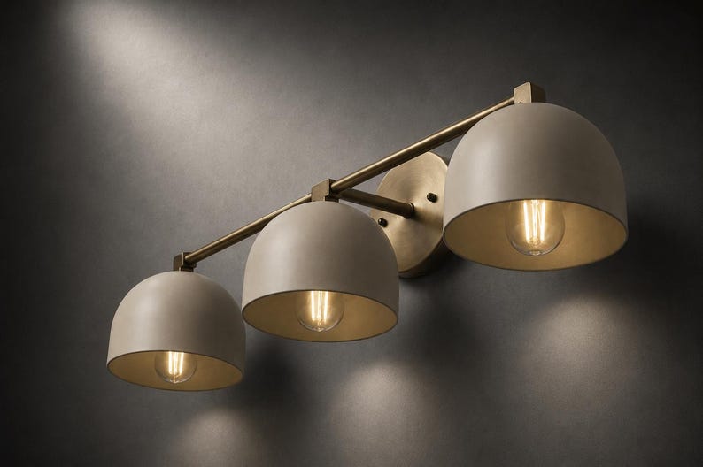May include: A three-light wall fitting featuring a horizontal brass bar and a circular wall mount. Three dome-shaped, matte beige shades are attached to the bar, each housing a single glowing filament light bulb that casts a warm light downwards.